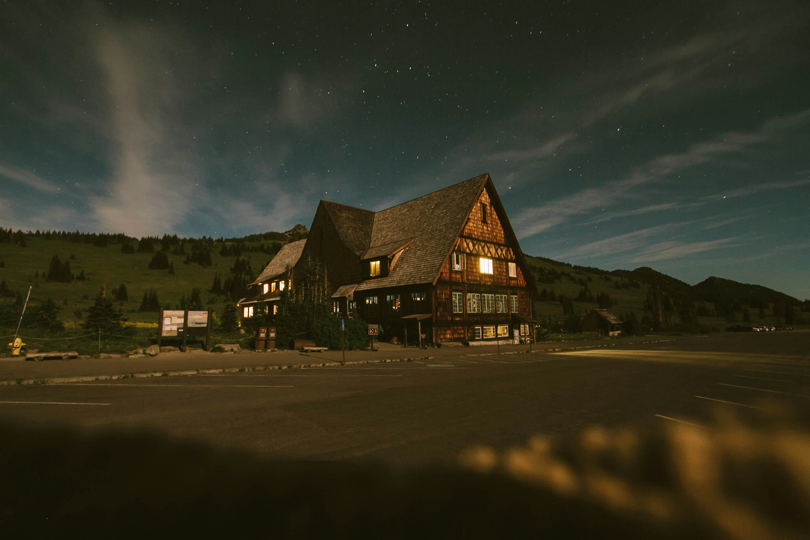 Large cabin-like house lit up from the inside at dusk in a field with mountains in the background